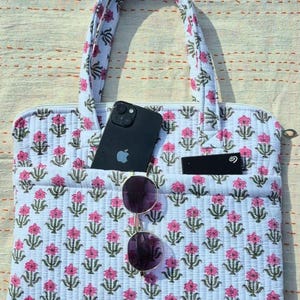 May include: A white quilted tote bag with a pink floral pattern. The bag has two pockets, one holding a black smartphone and the other a black card. A pair of sunglasses hangs from the front. The bag has a zipper closure and matching handles.