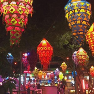 May include: A collection of tiered, multi-colored fabric lanterns hanging outdoors at night. The lanterns feature geometric patterns in shades of red, yellow, blue, and pink, illuminated from within to create a warm, glowing display.