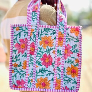 Puede incluir: Un bolso tote acolchado con base blanca y un patrón floral repetitivo en tonos rosa y naranja, acentuado por tallos y hojas verdes. El bolso incluye dos asas superiores y bordes ribeteados con una tela a rayas rosas y blancas.