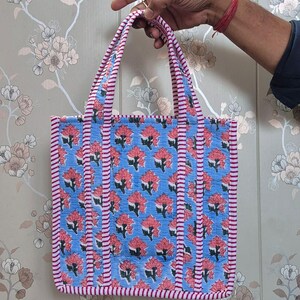 May include: A blue tote bag with a floral pattern in red and white. The bag has pink and white striped trim and handles. The bag is held up by a hand.