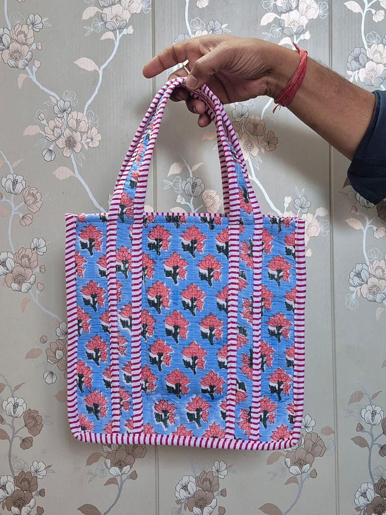 May include: A blue tote bag with a floral pattern in red and white. The bag has pink and white striped trim and handles. The bag is held up by a hand.