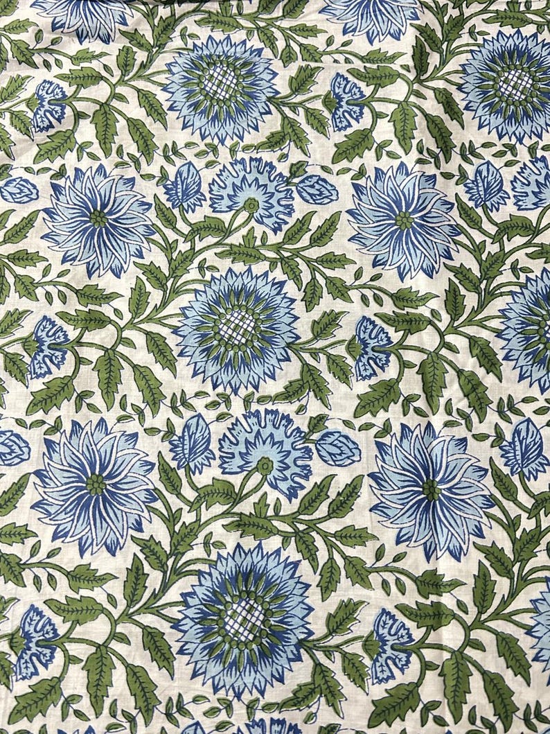 May include: Floral fabric with a repeating pattern of large blue flowers and green leaves on a cream background. The flowers have detailed petals and are surrounded by smaller buds and foliage. The design is symmetrical and creates a visually appealing textile.