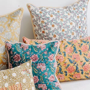 May include: A collection of square throw pillows featuring various floral block-print patterns in teal, mustard yellow, and cream. Each pillow is finished with a striped piping edge. One pillow displays the partial text "VA BIEN" in block letters.