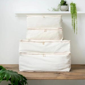 Puede incluir: Cuatro contenedores de almacenamiento blanquecinos apilados sobre una superficie de madera. Cada contenedor tiene un cierre de cremallera y una forma rectangular. Los contenedores son de diferentes tamaños, el más grande en la parte inferior y el más pequeño en la parte superior. Un estante con plantas está en el fondo.