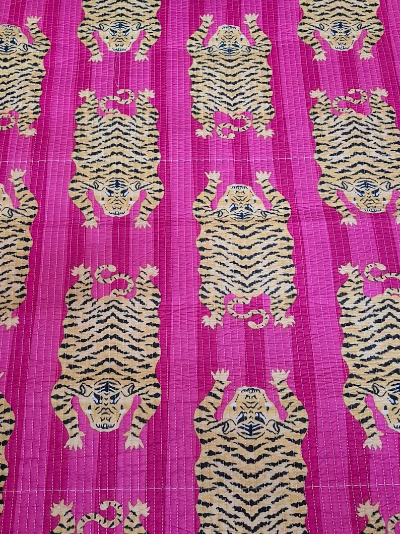 May include: A vibrant pink textile featuring a repeating pattern of stylized tigers. The tigers are depicted in tan and black stripes, with their paws raised. The background has vertical stripes, and the fabric appears to be quilted.