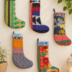 Op de afbeelding: Vijf gewatteerde kerstsokken gemaakt van kleurrijke patchworkstof met ingewikkelde kantha-stiksels. Elke sok heeft een unieke combinatie van patronen, waaronder bloemmotieven, geometrische vormen en strepen in levendige tinten groen, blauw, rood en geel.