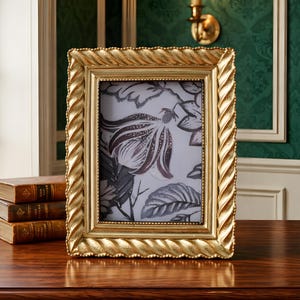Op de afbeelding: Een goudkleurige rechthoekige fotolijst met een gedraaide, touwachtige rand. De lijst bevat een zwart-witte bloemenprint. De lijst staat op een donker houten oppervlak, met boeken links.