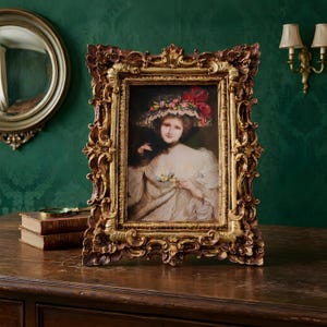 Pode incluir: Moldura dourada ornamentada com um retrato de uma mulher em vestido branco e chapéu floral. A moldura tem detalhes esculpidos e está sobre uma superfície de madeira.