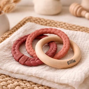 May include: Three baby teething rings in shades of red and natural wood, resting on a white cloth. One ring is engraved with the name "Ellie". The rings are made of silicone and wood. The background includes a woven mat and a vase with dried flowers.