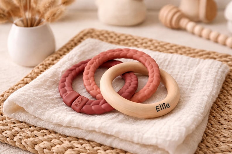 May include: Three baby teething rings in shades of red and natural wood, resting on a white cloth. One ring is engraved with the name "Ellie". The rings are made of silicone and wood. The background includes a woven mat and a vase with dried flowers.