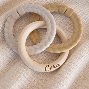 May include: Three baby teething rings. Two rings are made of a soft, textured material in light gray and tan colors. The third ring is made of natural wood and has the name "Cora" engraved on it. The rings are interlocked and rest on a cream-colored surface.