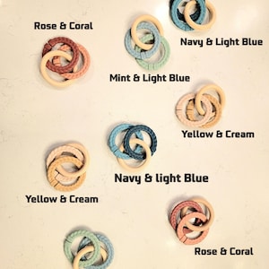 May include: Assortment of baby teething rings in various color combinations, including Rose & Coral, Navy & Light Blue, and Mint & Light Blue. Each ring features a braided texture and a natural wood-colored ring. The text labels each color combination.