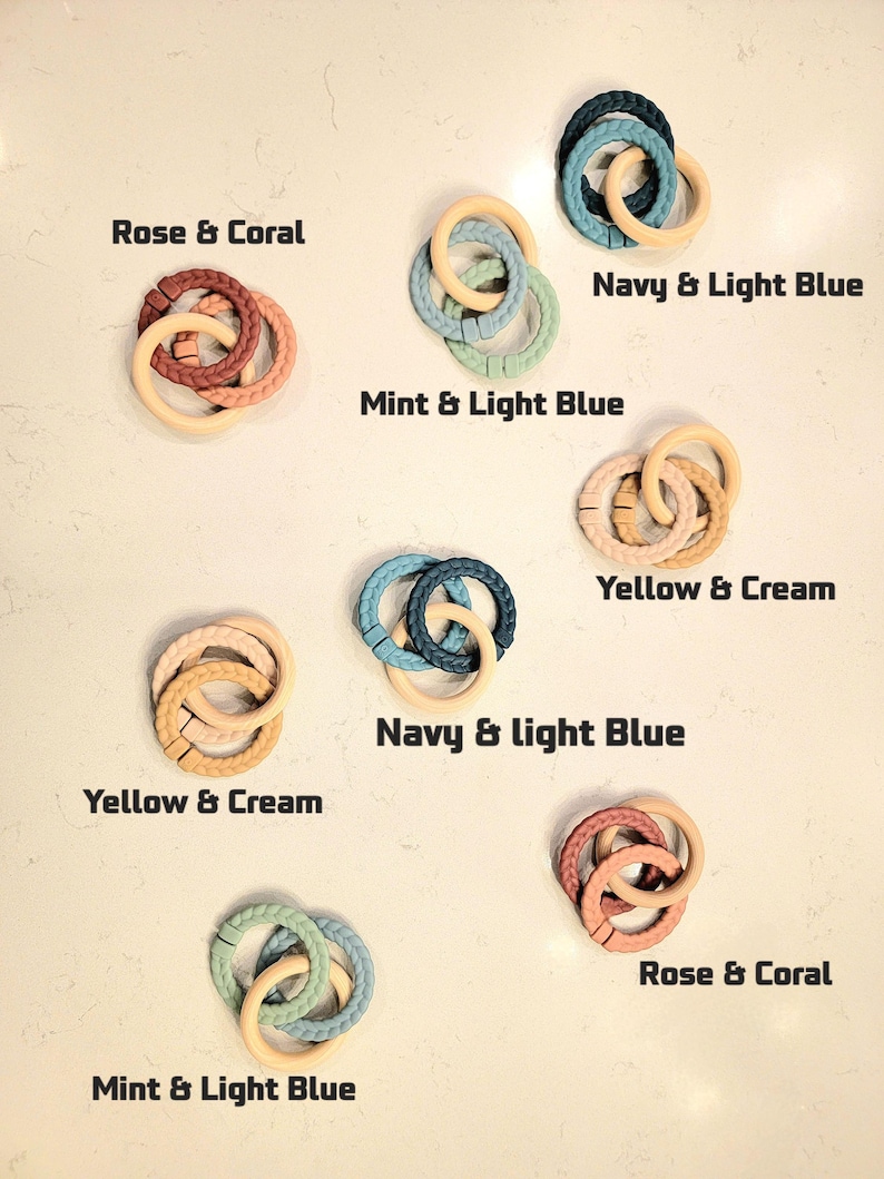 May include: Assortment of baby teething rings in various color combinations, including Rose & Coral, Navy & Light Blue, and Mint & Light Blue. Each ring features a braided texture and a natural wood-colored ring. The text labels each color combination.
