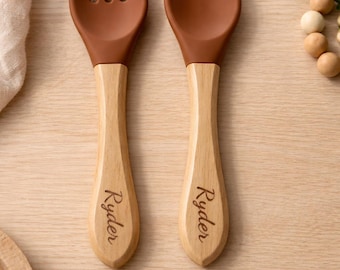 May include: A brown silicone fork and spoon set with wooden handles. The handles are engraved with the name "Ryder". The utensils are designed for babies or toddlers.