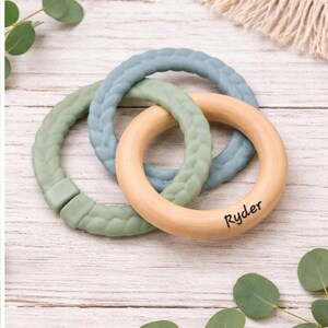 May include: A baby teething toy featuring three interlocking rings. Two rings are made of soft, textured silicone in shades of green and blue. The third ring is natural wood with the name "Ryder" printed on it.