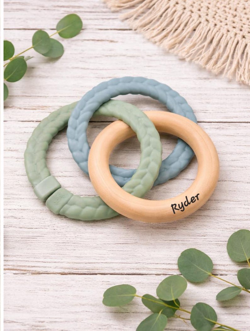 May include: A baby teething toy featuring three interlocking rings. Two rings are made of soft, textured silicone in shades of green and blue. The third ring is natural wood with the name "Ryder" printed on it.