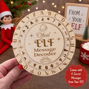 May include: A circular wooden message decoder toy featuring the engraved text Official ELF Message Decoder, a central Santa hat icon, and elf shoes. The outer ring displays the alphabet from A to Z alongside various holiday-themed icons.