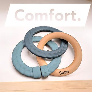 May include: Three interlocking teething rings in shades of blue and natural wood. The text "Comfort" is displayed above the rings, and the name "Sean" is printed on one ring. The text "Did you know? Teething can start as early as 4 months old!" is also visible.