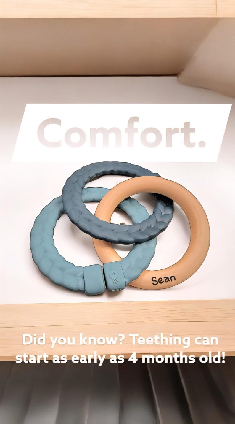 May include: Three interlocking teething rings in shades of blue and natural wood. The text "Comfort" is displayed above the rings, and the name "Sean" is printed on one ring. The text "Did you know? Teething can start as early as 4 months old!" is also visible.