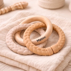 May include: Three wooden baby teething rings in neutral colors, one with the name "Mason" printed on it. The rings are on a folded, textured, off-white cloth. A wooden teething toy is in the background.