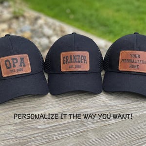 May include: Three black trucker hats with mesh backs and solid front panels. Each hat features a rectangular brown leather patch. The patches are engraved with the text OPA EST. 2026, GRANDPA EST. 2026, and YOUR PERSONALIZATION HERE.