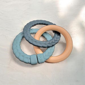 May include: A baby teether toy featuring three interlocking rings. Two rings are a textured blue-green silicone, and one is a smooth, light-colored wood. The rings are designed to be grasped and chewed by infants.