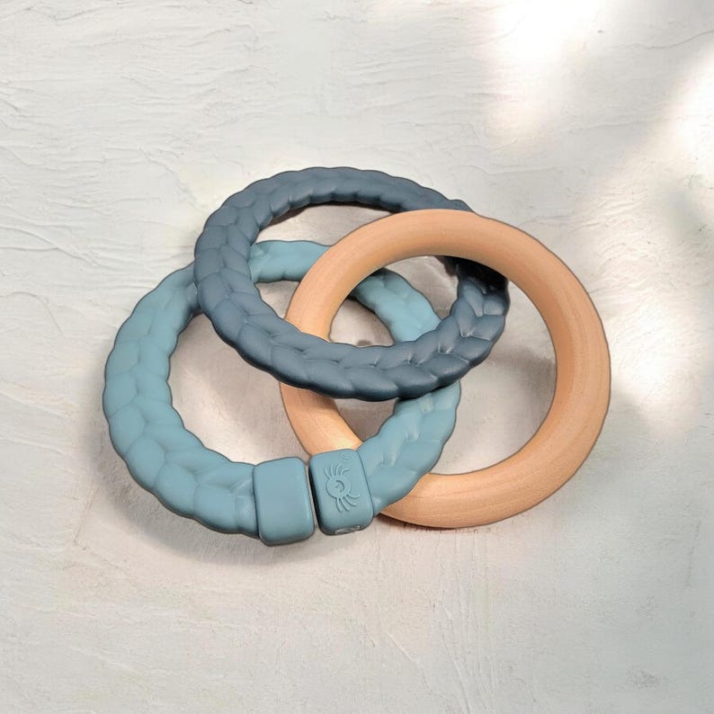 May include: A baby teether toy featuring three interlocking rings. Two rings are a textured blue-green silicone, and one is a smooth, light-colored wood. The rings are designed to be grasped and chewed by infants.