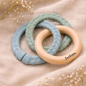 May include: Three interlocking baby teething rings. Two are a braided design in blue and green, and one is natural wood with the name "Jackson" printed on it. The rings are on a beige fabric background.