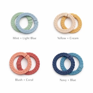 May include: Four pairs of braided silicone teething rings in various color combinations: mint and light blue, yellow and cream, blush and coral, and navy and blue. Each pair is connected by a small, color-matched clasp. Text labels identify each color pairing.