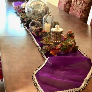 May include: A decorative table runner in deep purple with a silver and black beaded trim. The runner is adorned with fall-themed decorations, including pumpkins, a large glass vase, and a candle in a decorative cage. The table is made of wood.