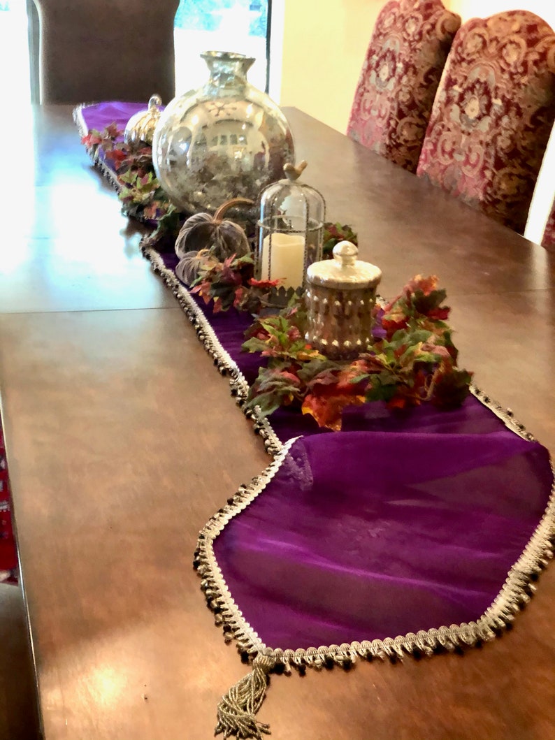 May include: A decorative table runner in deep purple with a silver and black beaded trim. The runner is adorned with fall-themed decorations, including pumpkins, a large glass vase, and a candle in a decorative cage. The table is made of wood.