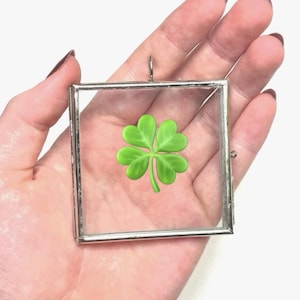 May include: A silver-toned metal picture frame with a glass front, holding a pressed four-leaf clover. The frame has a small loop at the top for hanging.