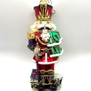 May include: A decorative Christmas ornament shaped like a nutcracker soldier. The ornament features a red and gold uniform, a white beard and hair, and a black mustache. It holds a sword and a green cape.