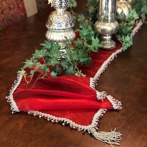 May include: A red table runner with a silver beaded trim and tassel. The runner is decorated with silver candle holders and faux greenery. The table is made of dark wood.