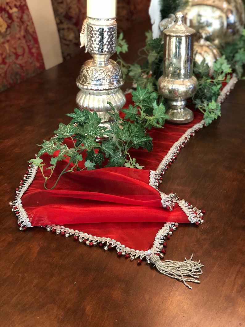 May include: A red table runner with a silver beaded trim and tassel. The runner is decorated with silver candle holders and faux greenery. The table is made of dark wood.