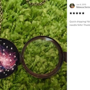 May include: A round, silver locket necklace with a pink and blue galaxy design inside. The locket is open and the inside is visible. The locket is on a green fuzzy surface.