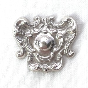 May include: A silver-toned metal ornament with a decorative, ornate design. The ornament has a raised, round center and intricate scrollwork around the edges.