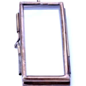 May include: A small, rectangular, brass picture frame with a glass front and a brass loop at the top for hanging. The frame is designed to hold a small photograph or other memento.