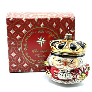 May include: A glass Christmas ornament shaped like a nutcracker soldier, wearing a gold crown and red jacket, with a candy cane. The ornament is next to a red box with gold star patterns and the text "Christopher Radko".