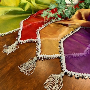 May include: A collection of colorful, sheer table runners in shades of green, red, gold, and purple. Each runner has a beaded trim and a tassel at the end. The runners are arranged on a wooden table.
