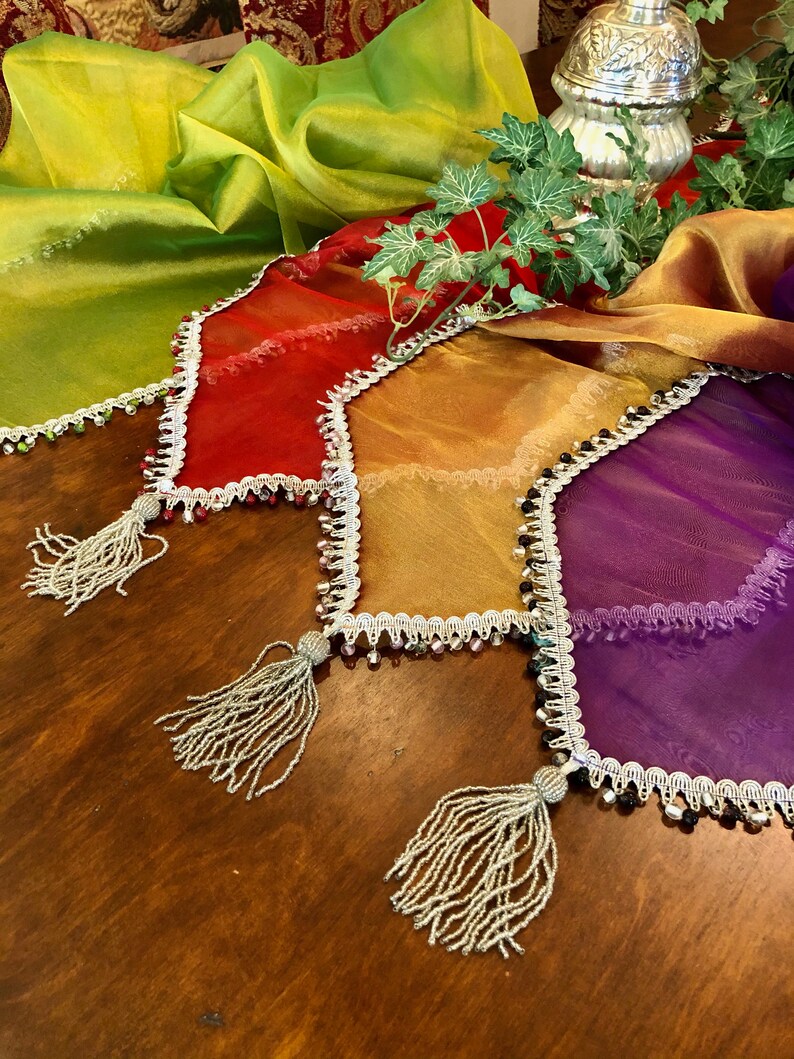 May include: A collection of colorful, sheer table runners in shades of green, red, gold, and purple. Each runner has a beaded trim and a tassel at the end. The runners are arranged on a wooden table.