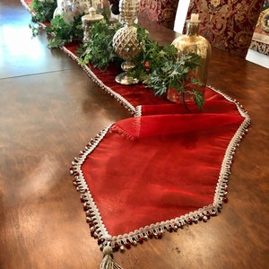 May include: A red sheer table runner with a white and silver beaded trim and a beaded tassel. The runner is decorated with silver and gold decorative items, greenery, and candles, creating a festive holiday theme.