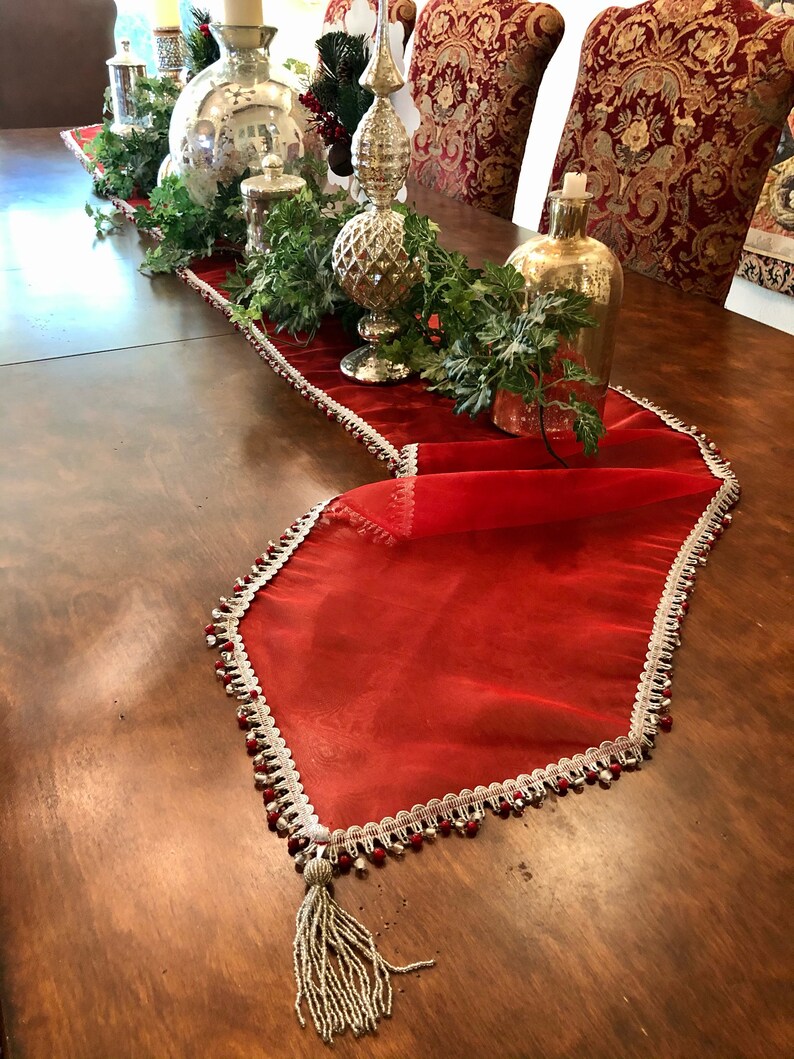 May include: A red sheer table runner with a white and silver beaded trim and a beaded tassel. The runner is decorated with silver and gold decorative items, greenery, and candles, creating a festive holiday theme.