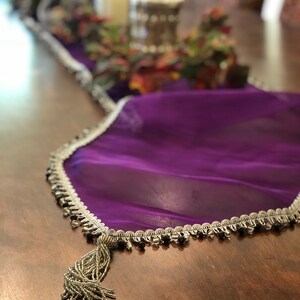 May include: A purple sheer table runner with a silver beaded trim and a beaded tassel. The runner is draped over a wooden table, with decorative items in the background.