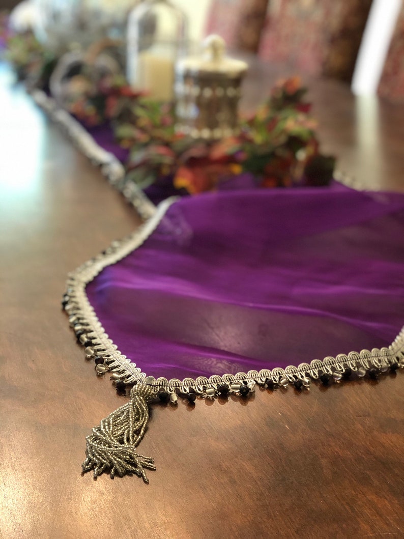 May include: A purple sheer table runner with a silver beaded trim and a beaded tassel. The runner is draped over a wooden table, with decorative items in the background.