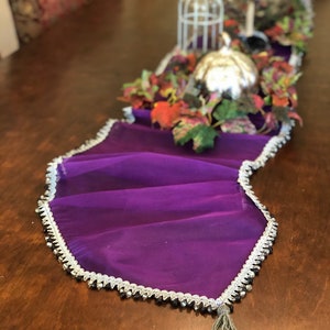 May include: A vibrant purple table runner with a silver beaded trim and a gray tassel. The runner is draped on a wooden table, with decorative silver spheres, a small birdcage, and fall foliage accents.
