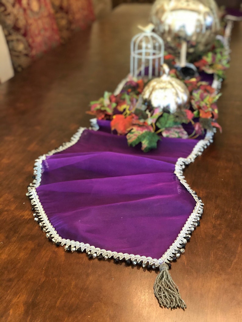 May include: A vibrant purple table runner with a silver beaded trim and a gray tassel. The runner is draped on a wooden table, with decorative silver spheres, a small birdcage, and fall foliage accents.