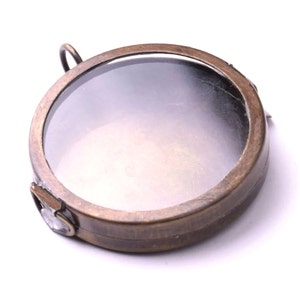May include: A round, brass-colored magnifying glass with a clear glass lens. The magnifying glass has a hinged lid and a small loop at the top for hanging.