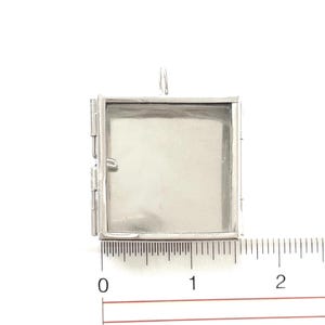 Może przedstawiać: Srebrny metalowy medalion z otwieranym zawiasem. Medalion ma około 2,5 cm szerokości i jest pokazany obok linijki.