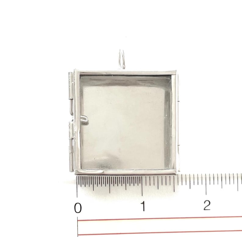 Może przedstawiać: Srebrny metalowy medalion z otwieranym zawiasem. Medalion ma około 2,5 cm szerokości i jest pokazany obok linijki.